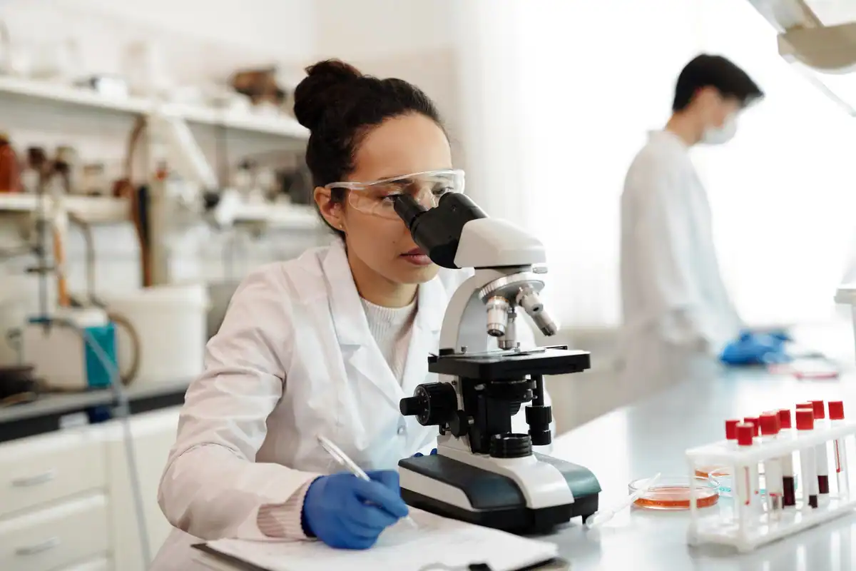 Cercetătoare în halat alb privește printr-un microscop într-un laborator luminos