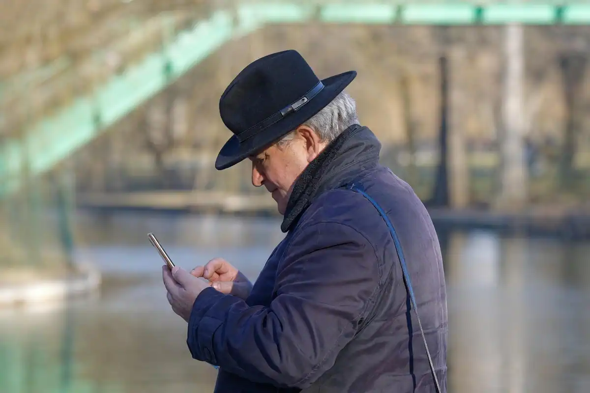 Bărbat în vârstă, cu pălărie, se uită la telefon într-un parc