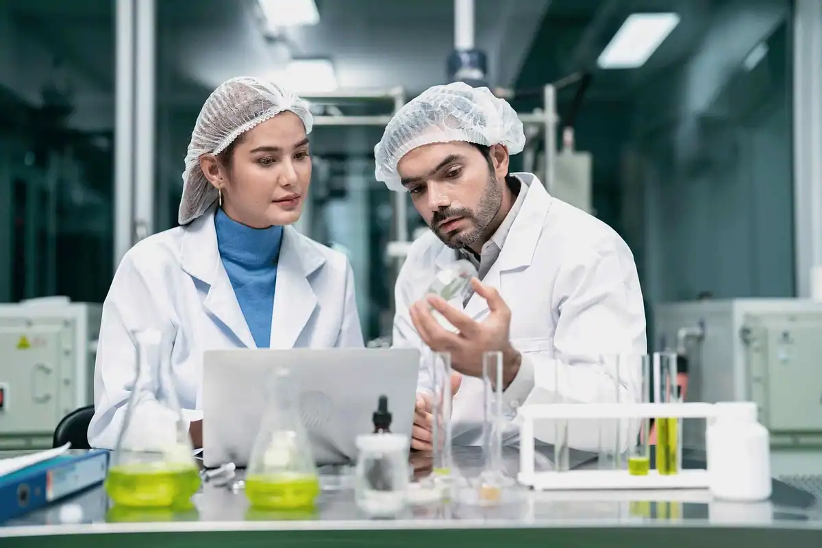 Doi cercetători în halate și bonete discută într-un laborator curat